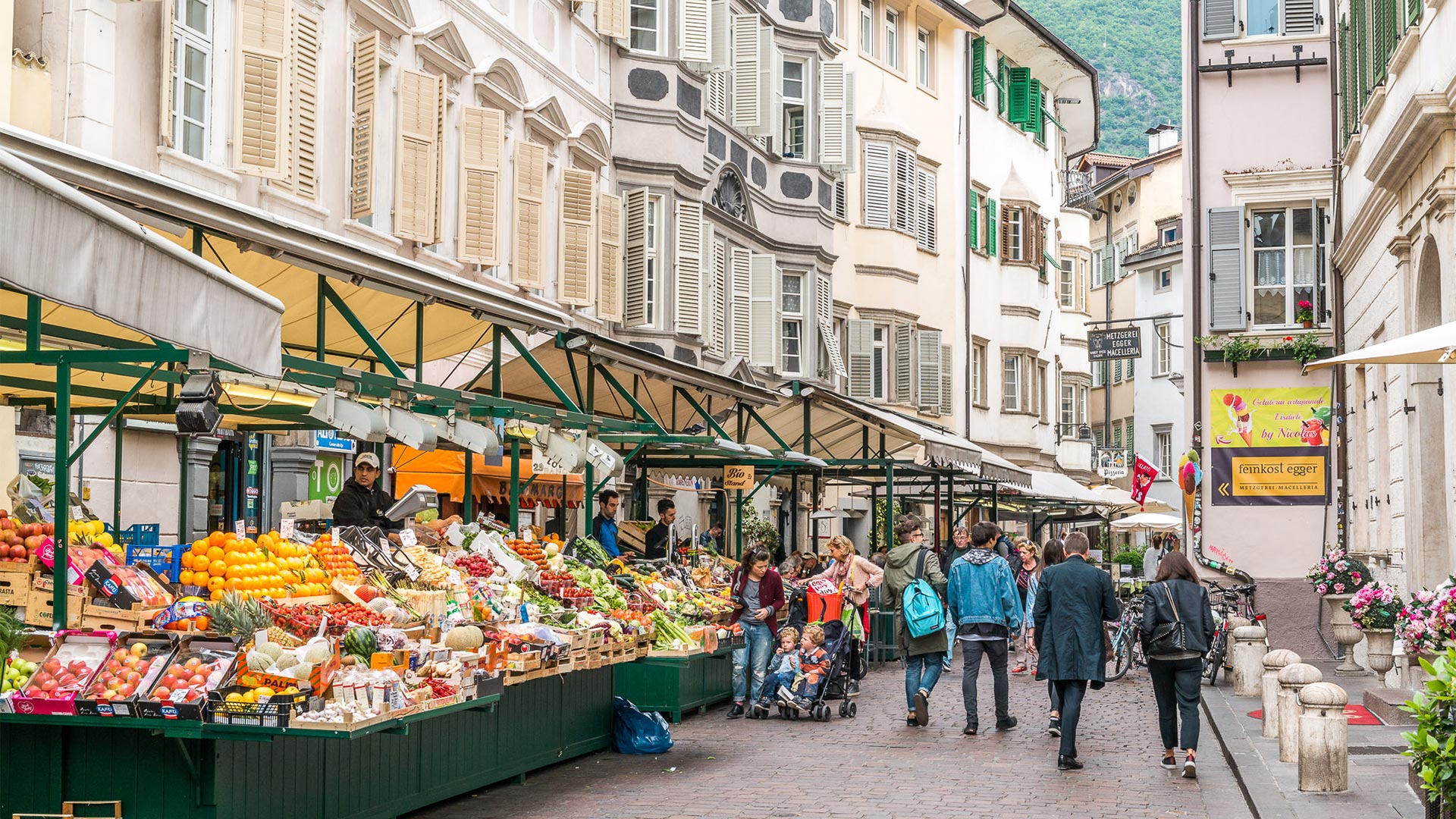 In Piazza Erbe i cittadini e turisti si fermano ai mercatini e fruttivendoli per comprare prodotti locali freschi.